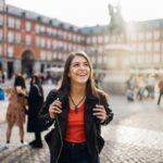 A young female tourist holding her backpack's straps and smiling in the Plaza Mayor in Madrid, Spain.