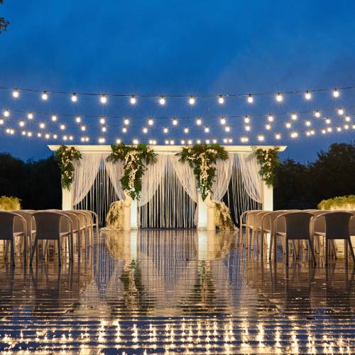 A wedding canopy is empty and brightly lit at night in front of a crowd of chairs while string lights hang above.