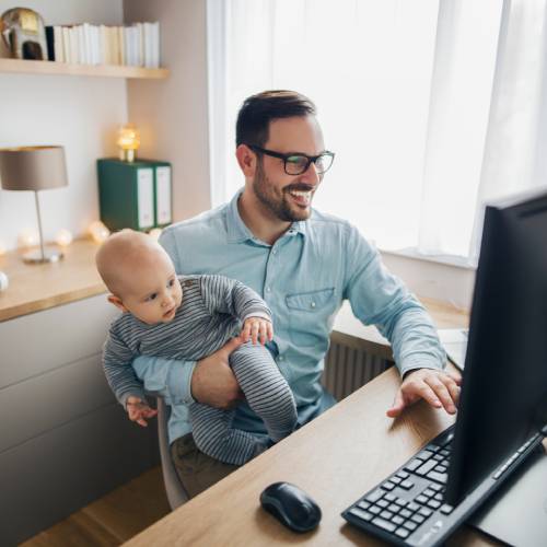 A father with his infant son on his lap is working remotely on a laptop inside his family’s home office.