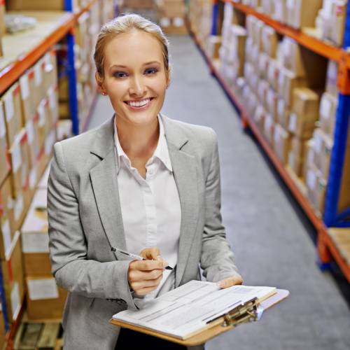 A woman in business attire stands holding a clipboard and pen in a storage warehouse full of cardboard boxes.