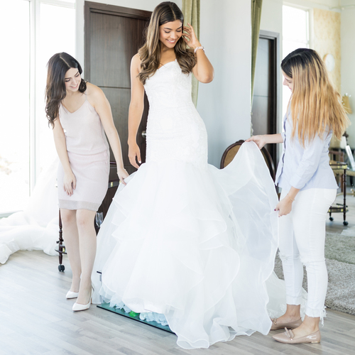 A woman stands on a stool while two stylists fix her wedding dress. The woman looks down at the bottom of the dress.