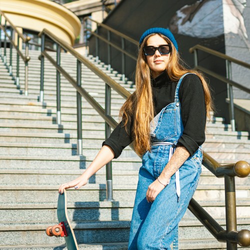 A woman on the steps of a building with her skateboard. She's wearing a denim jumpsuit and a black beanie.