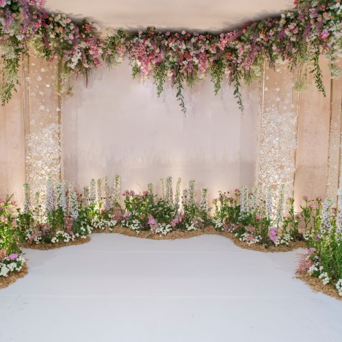 A wedding backdrop with hanging flowers and decorative plants. There are beams of light illuminating the ground.