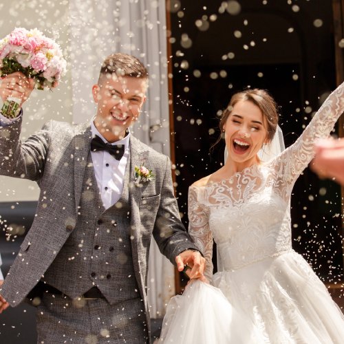 A bride and groom smile while holding hands and walking out of a church. Their loved ones throw rice at them.