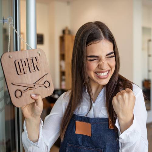 A woman excitedly turned the closed sign to open for her haircutting business. She is wearing a pair of overalls.