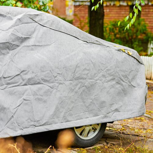 The back end of a car is covered with a light gray vehicle cover to protect it from the rain and falling leaves.