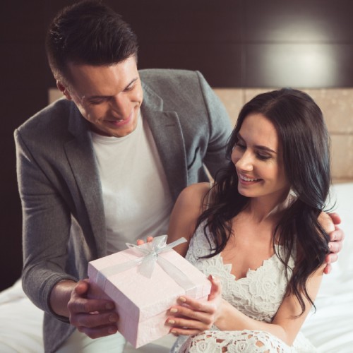 Newlyweds sitting together on a bed as they smile at a gift. The gift box is medium-sized and the wrap is white with a bow.