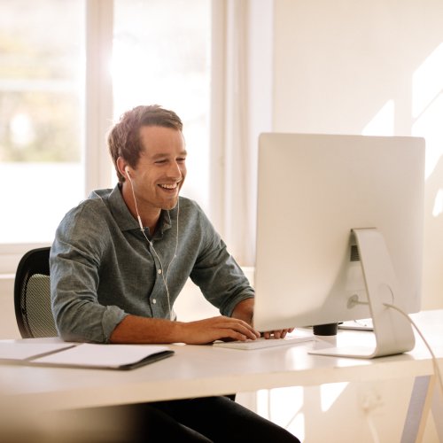 A man sitting at a long white desk with a keyboard and a monitor. He's smiling while wearing wired earbuds.