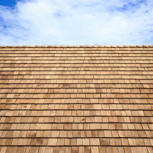 A roof with wooden shingles in various shades of brown. There is a blue sky in the background with a puffy white cloud.