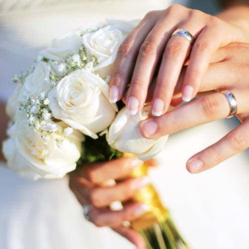 A husband and wife wearing wedding attire both place their hands over each other on the wife's white bouquet.