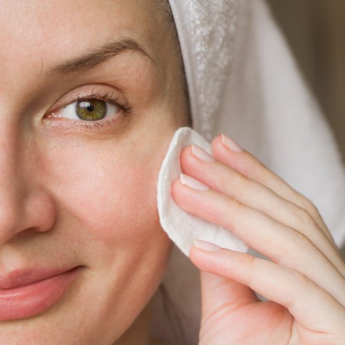 A close-up of a smiling woman with a white towel on her head using a white cotton pad to wipe her face.