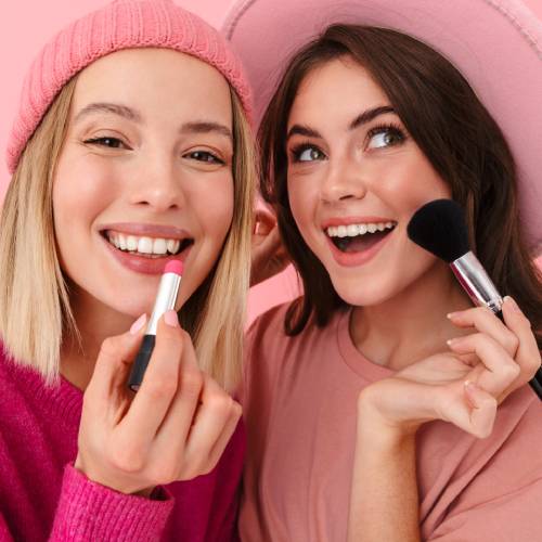 Two smiling women are applying makeup to their faces. They are both wearing pink shirts and pink hats.