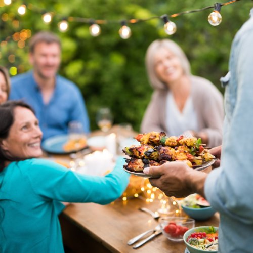 A person carrying skewers on a plate to an outdoor table adorned with decorations and food surrounded by seated people.