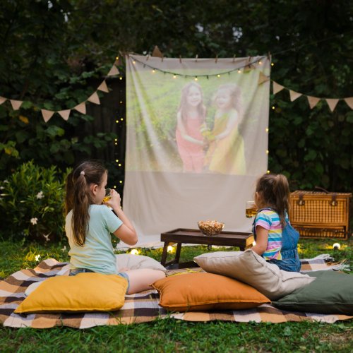 Two small children sitting in a designated play area in the backyard. They have a projector screen, pillows, and snacks.