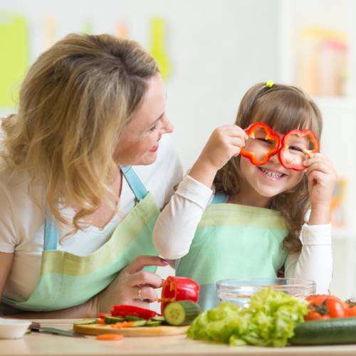 A woman and a young girl smile in the kitchen. The girl holds slices of red bell pepper in front of her eyes.