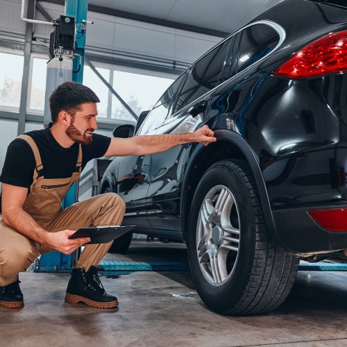 A young man in overalls holding a clipboard while crouching down to examine the tires on a black midsized car.