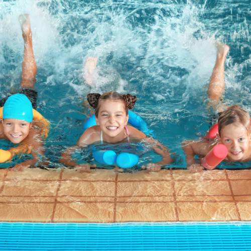 Three children swimming in a pool. They each have pool noodles as they splash their legs in the water.