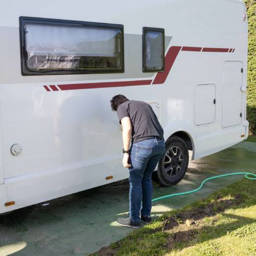 A person in a black shirt and jeans bends over to inspect a white, parked RV. The driver's side door is open on the RV.