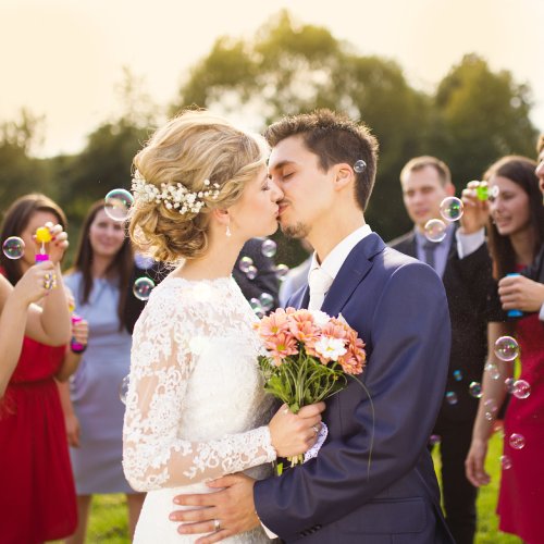 A bride and groom sharing a kiss outside while their wedding guests blow bubbles in the background.