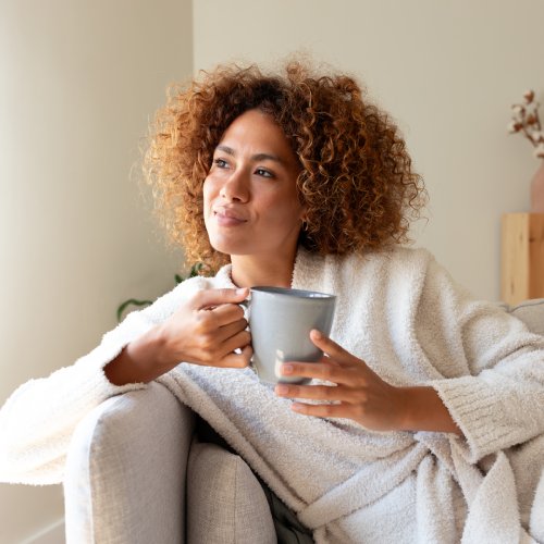 A woman in a robe is holding a mug, smiling gently, leaning against a sofa's armrest, and looking toward some light.