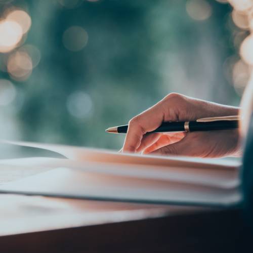 A close-up of a woman's hand holding a pen over a notebook at a wooden desk with the background blurred.