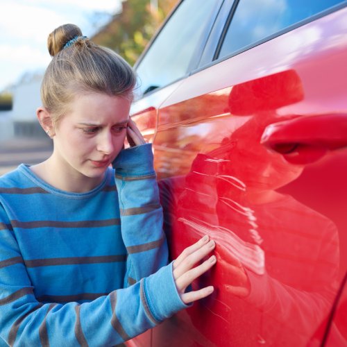 A teenage girl is inspecting scratches on a red car. She has a worried expression as she touches the scratches.