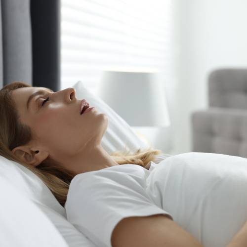 A woman asleep in her bed with her arms folded on her stomach. Her mouth is open slightly as she snores.