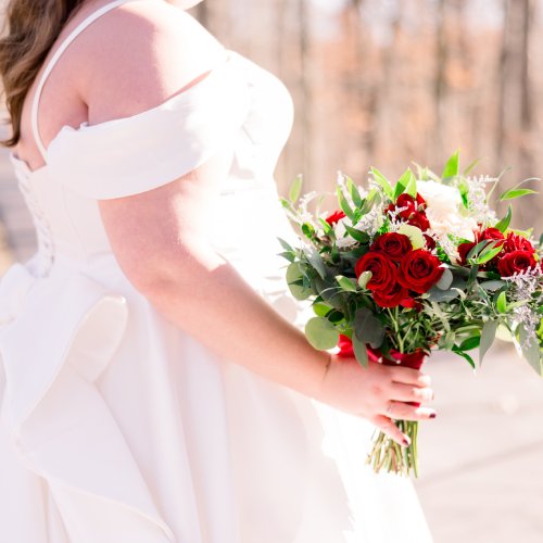 A plus-size bride in her satiny gown. She's holding a bouquet with red roses and standing on an outdoor path lined with trees.