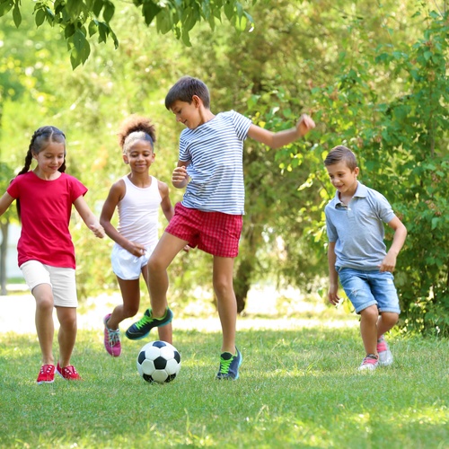 Two young boys and two young girls are outside playing. One of the young boys is kicking a soccer ball.