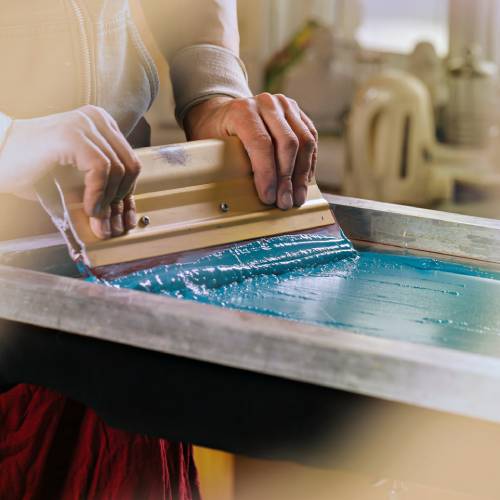 A pair of hands running a squeegee down a frame for screen printing T-shirts. The frame is full of bright blue ink.