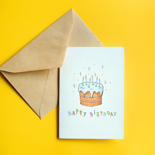 A birthday card with an illustration of a cake with candles. The card is on top of a brown envelope on a bright yellow background.