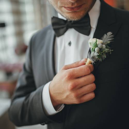 A groom in a black suit with a matching bowtie adjusting the boutonniere pinned to his left notched lapel.