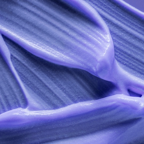 A close-up view of thick purple shampoo spread across a surface. The product is glossy with ridged and creamy patterns.