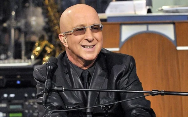 Paul Shaffer smiling while seated at a piano microphone, wearing a black suit and sunglasses during a live performance