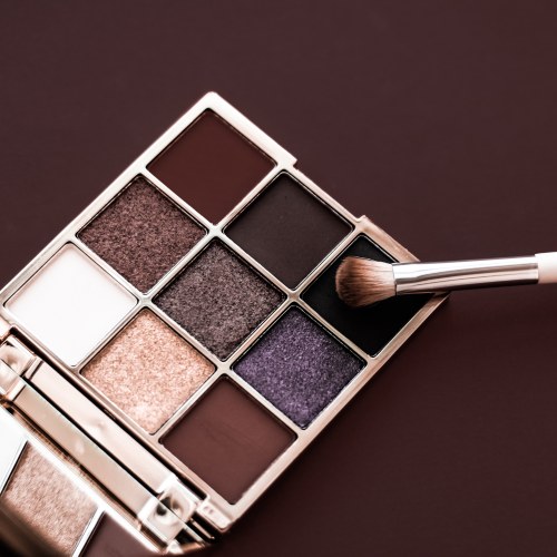 Eyeshadow palette with makeup brush applying dark shade on professional cosmetic kit against a brown background.