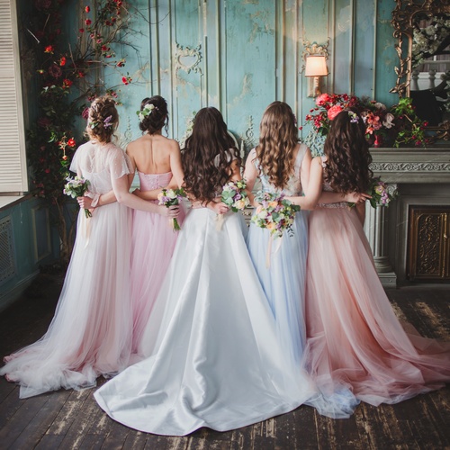 A bride and bridesmaids stand together indoors, facing a wall, wearing formal dresses in soft colors near a decorated wall.