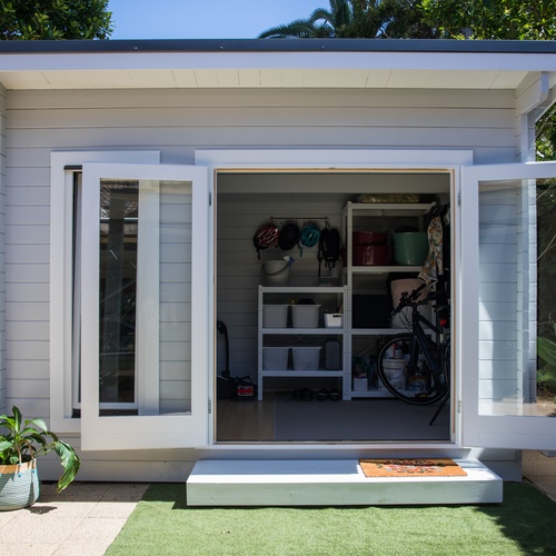 White backyard shed with doors open, showing organized shelves, boxes, and a bicycle parked inside.