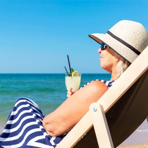A woman in a blue and white striped dress and hat sits on a beach lounger by the sea, holding a drink and smiling.