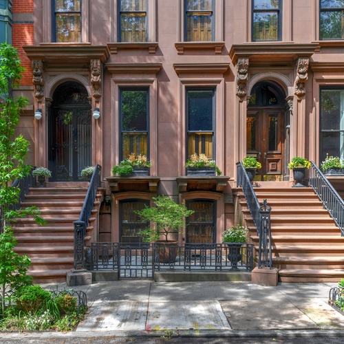 Row of brownstone townhouses with stoops, iron railings, and potted plants along a tree-lined sidewalk.