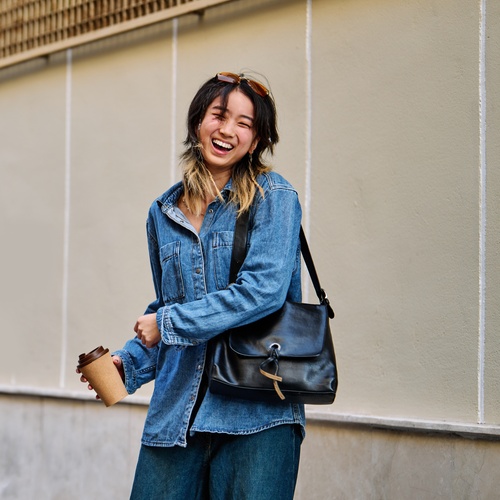 A smiling woman holding a takeaway cup while standing on a sunlit city street with buildings behind her.