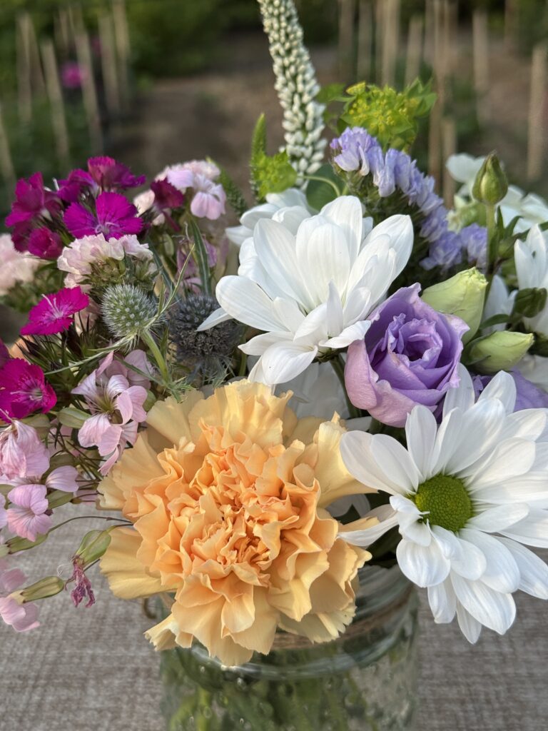 Fresh summer flower arrangement in a mason jar with white daisies, lavender roses, peach carnations, and wildflowers at an outdoor dining table in Westchester, NY