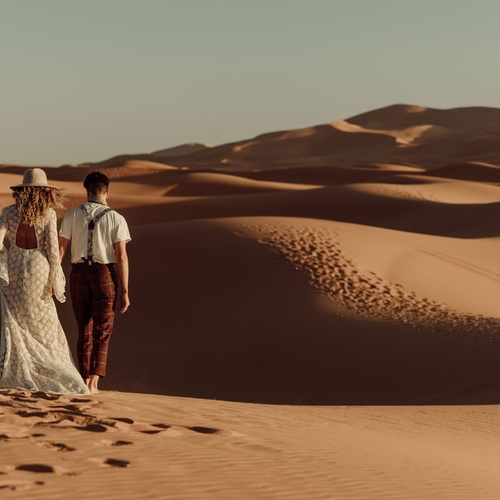 A woman wearing a sunhat and a wedding dress holds hands with a man wearing suspenders. They walk through sand dunes.