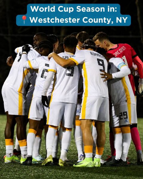 A nighttime soccer team huddles together on a turf field in Westchester County, New York. Players in white uniforms with yellow accents gather in a tight circle before a match, while a goalkeeper in a bright pink jersey stands at the edge of the group. A blue banner at the top of the image reads, “World Cup Season in: Westchester County, NY,” with a red location pin icon. The scene conveys teamwork, anticipation, and the excitement of soccer season.