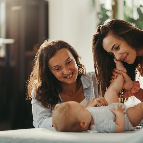 A baby lies on a bed while two smiling women lean close, one holding and kissing the baby's raised foot.