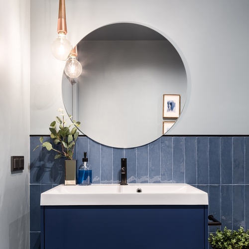 A bathroom with ivory walls, a vertical blue backsplash, a round mirror, and a dark blue vanity with a white counter.