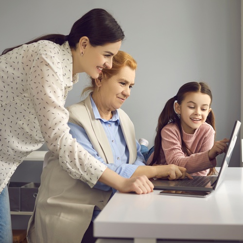 A woman, an older woman, and a young girl smile together while using a laptop beside a sunny window.