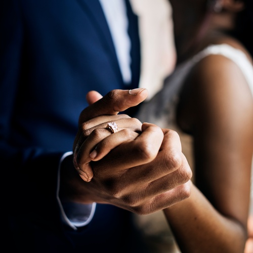 A bride and groom holding hands on their wedding day, with a ring visible and the groom wearing a dark suit.