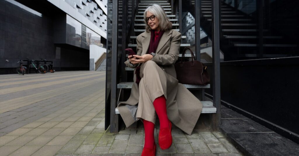 A stylish older woman in a tan coat and red boots sits on outdoor stairs, looking at her phone in a modern city setting.