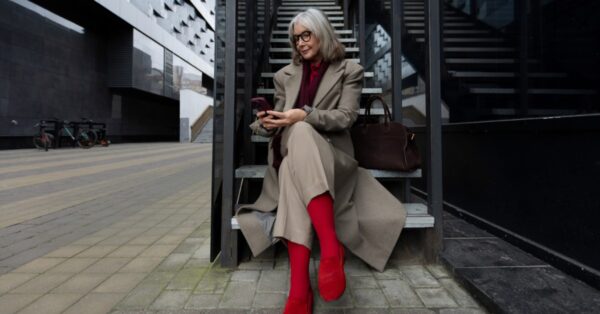 A stylish older woman in a tan coat and red boots sits on outdoor stairs, looking at her phone in a modern city setting.