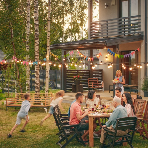 Adults and children gathered around an outdoor table with food, string lights, colorful flags, trees, and a house.